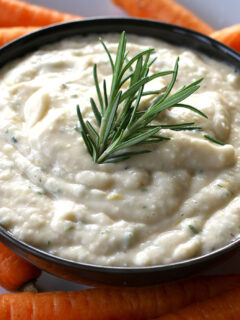 A bowl of white bean dip with carrots and a sprig of rosemary.
