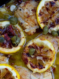A dish of lemon chicken piccata with olives on a wooden table.