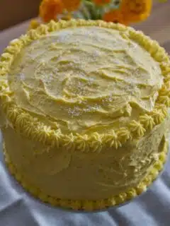 A classic yellow cake is sitting on a table in front of flowers, homemade buttercream frosting.